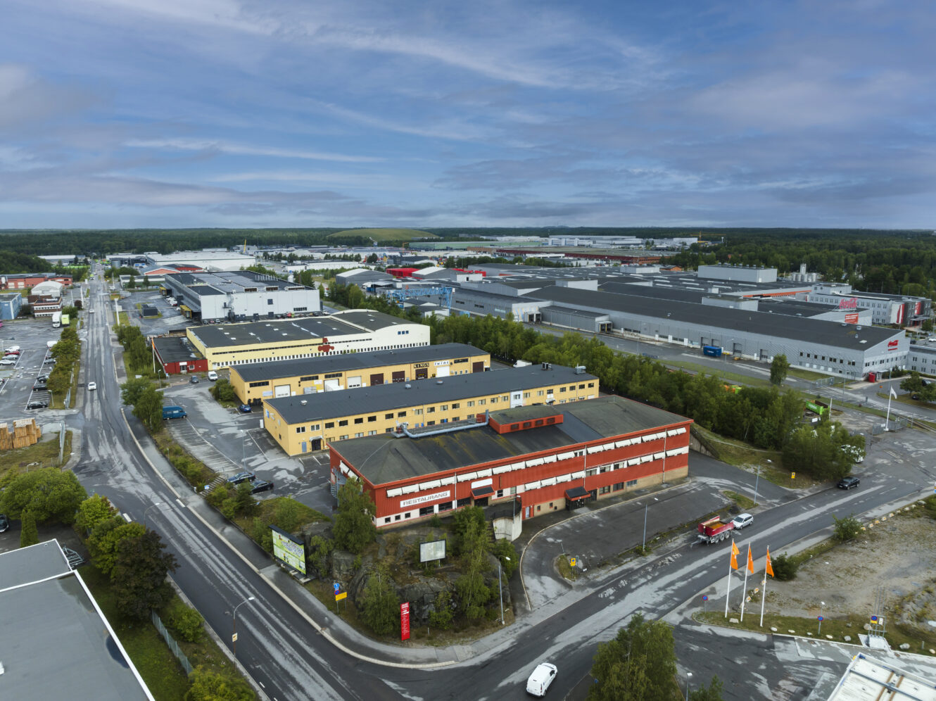 Flygfoto över ett industriområde med stora lagerlokaler, kontorsbyggnader, parkeringsplatser och omgivande vägar, med en skogsklädd bakgrund och molnig himmel.