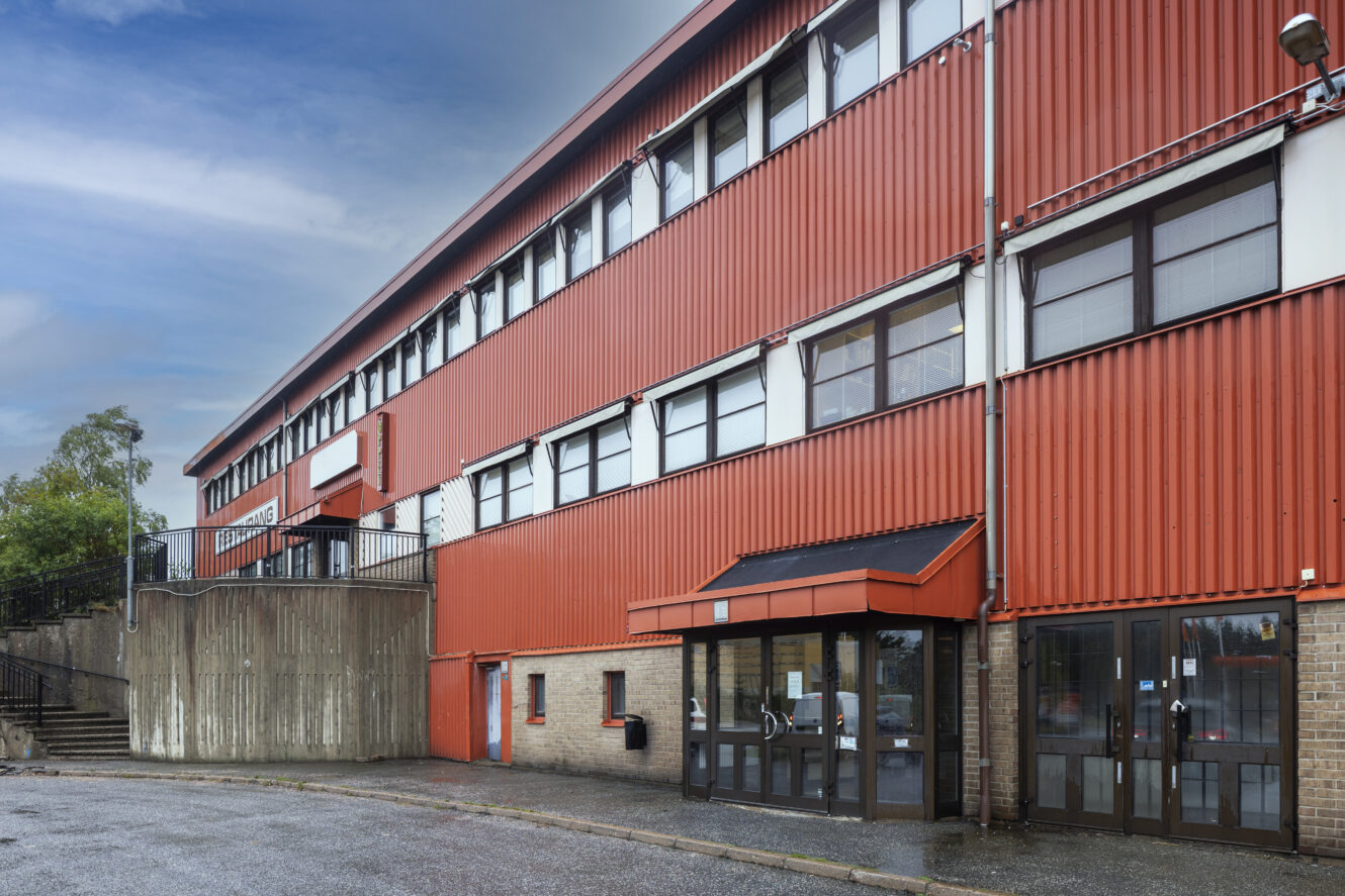 En trevåningsbyggnad med röd metallfasad och många fönster, med en ingångsdörr i glas och betongtrappa på vänster sida.