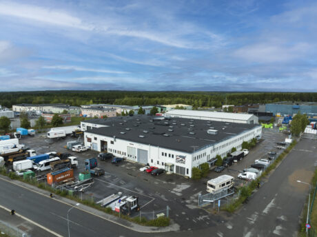 Ett stort industriellt lager med lastbilar, skåpbilar och bilar parkerade utanför, omgivet av en inhägnad tomt i ett kommersiellt område under en blå himmel.