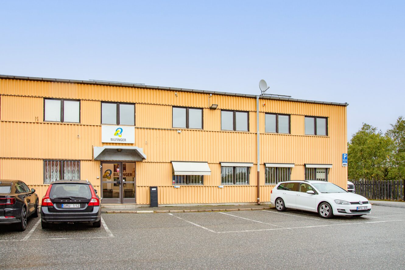 En gul tvåvåningsbyggnad med flera fönster, en skylt ovanför entrén och parkerade bilar framför en mulen dag.