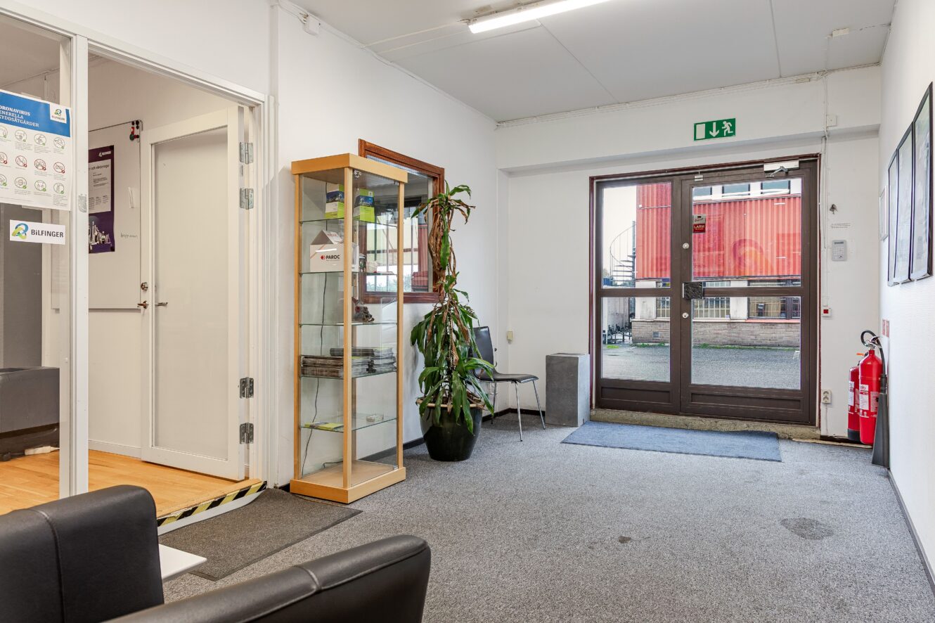 Kontorslobby med grå matta, vitrinskåp, krukväxt, brandsläckare, stolar och en utgångsdörr i glas som leder ut till ett industriområde.