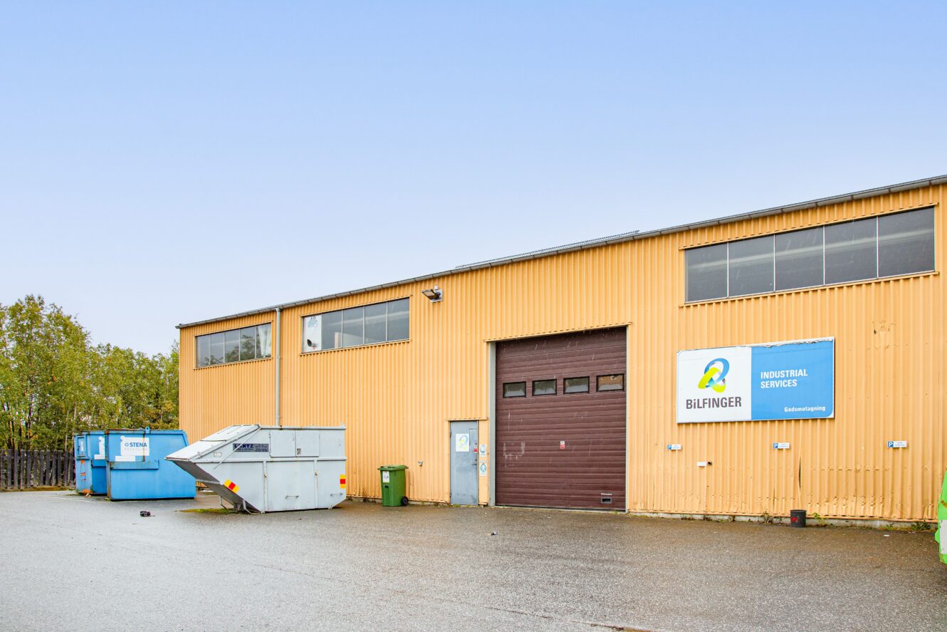 En gul industribyggnad med en stor garageport, avfallsbehållare och en skylt med texten Bilfinger Industrial Services på ytterväggen.