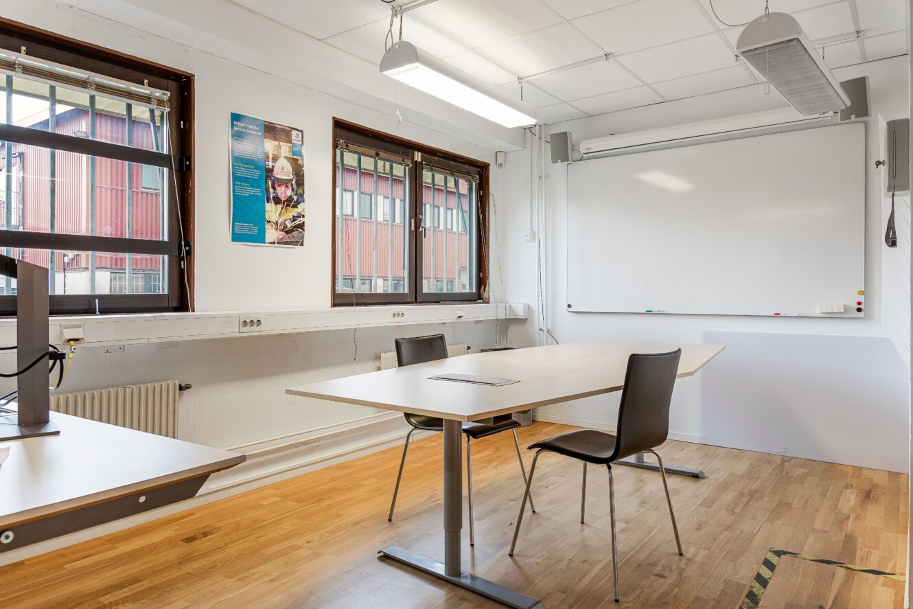 Ett klassrum med två bord, tre stolar, en whiteboard, fönster med spröjs och en affisch på väggen. Rummet har trägolv och takbelysning.