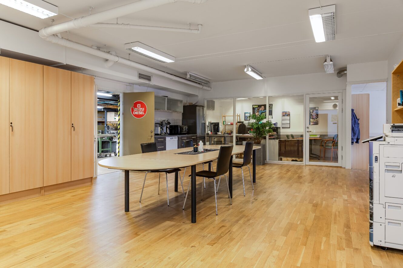 Ett rymligt kontorsområde med ett träbord och stolar i förgrunden, förvaringsskåp till vänster, en kopiator till höger och kontor och utrustning med glasväggar i bakgrunden.