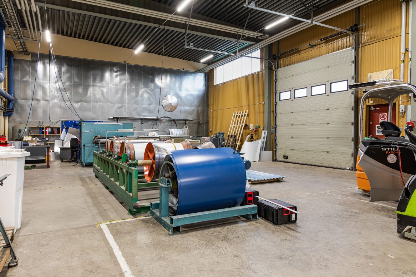 Industriverkstad med stora metallrullar på ett grönt ställ, en rullport, diverse utrustning och takbelysning.