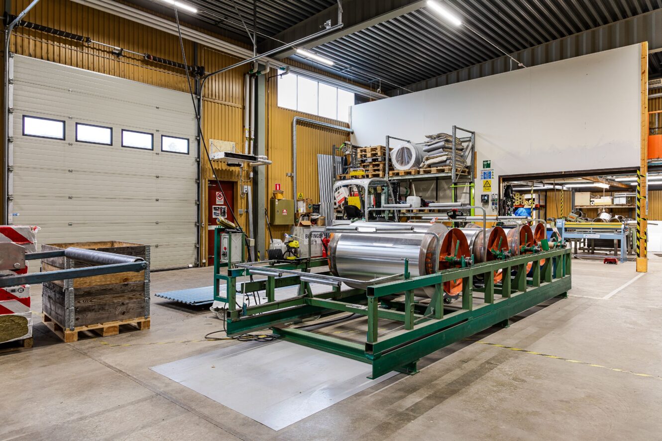 Industriell verkstadsinredning med stora metallrullar på en grön maskin, lastpallar och diverse utrustning; öppen garageport syns i bakgrunden.