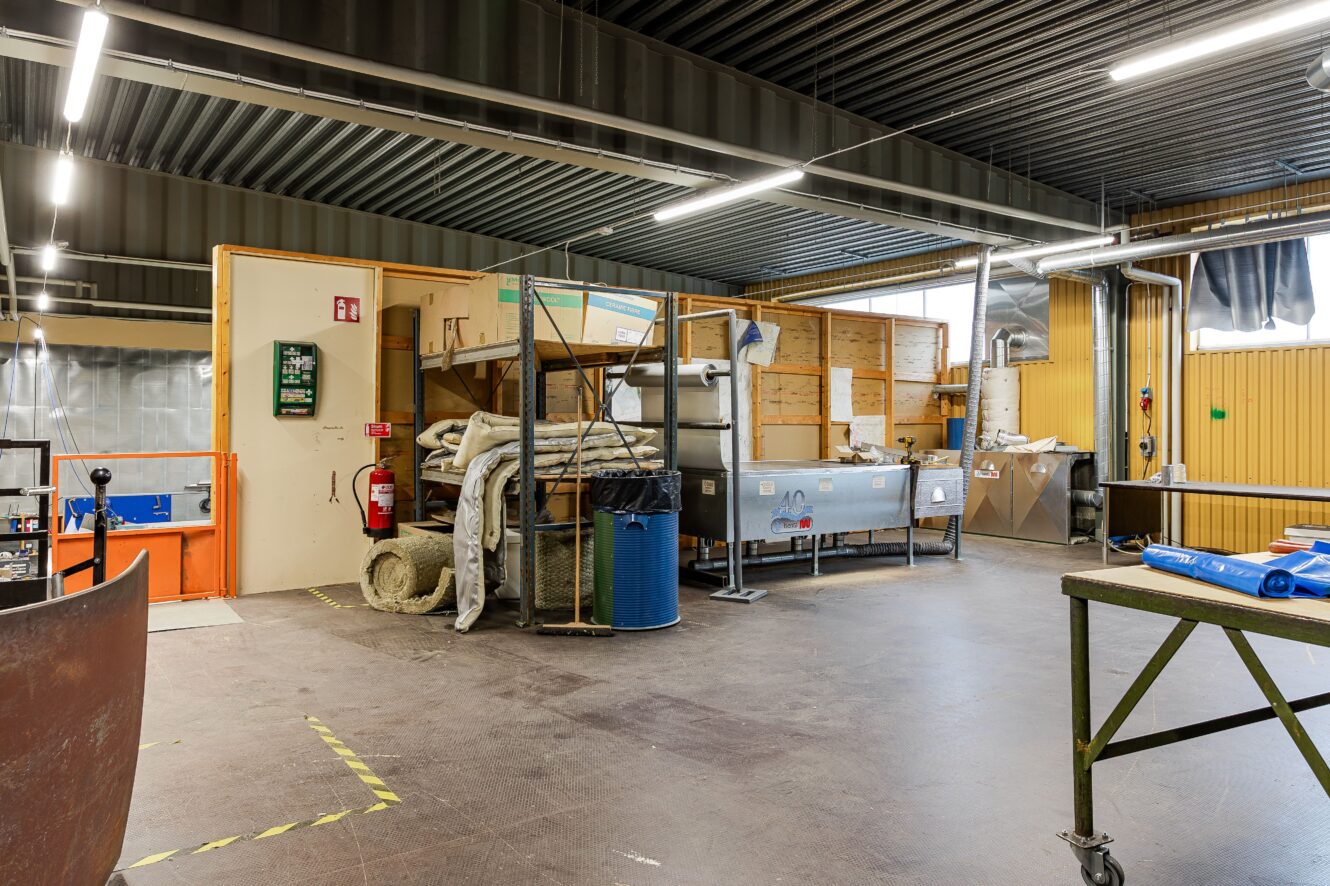 Industriellt arbetsutrymme med diverse utrustning, isoleringsmaterial, metallbord och förvaringshyllor. Det finns lysrörsbelysning och synliga ventilationskanaler.