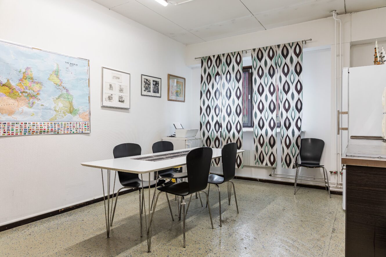 Ett litet mötesrum med ett vitt bord, fem svarta stolar, en världskarta på väggen, inramade tavlor och mönstrade gardiner för ett fönster.