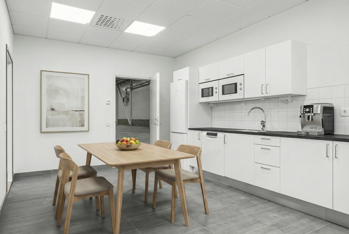 Modernt kök med vita skåp, ett matbord i trä med fyra stolar, en skål med frukt, inbyggda vitvaror och en kaffemaskin på ett grått klinkergolv.