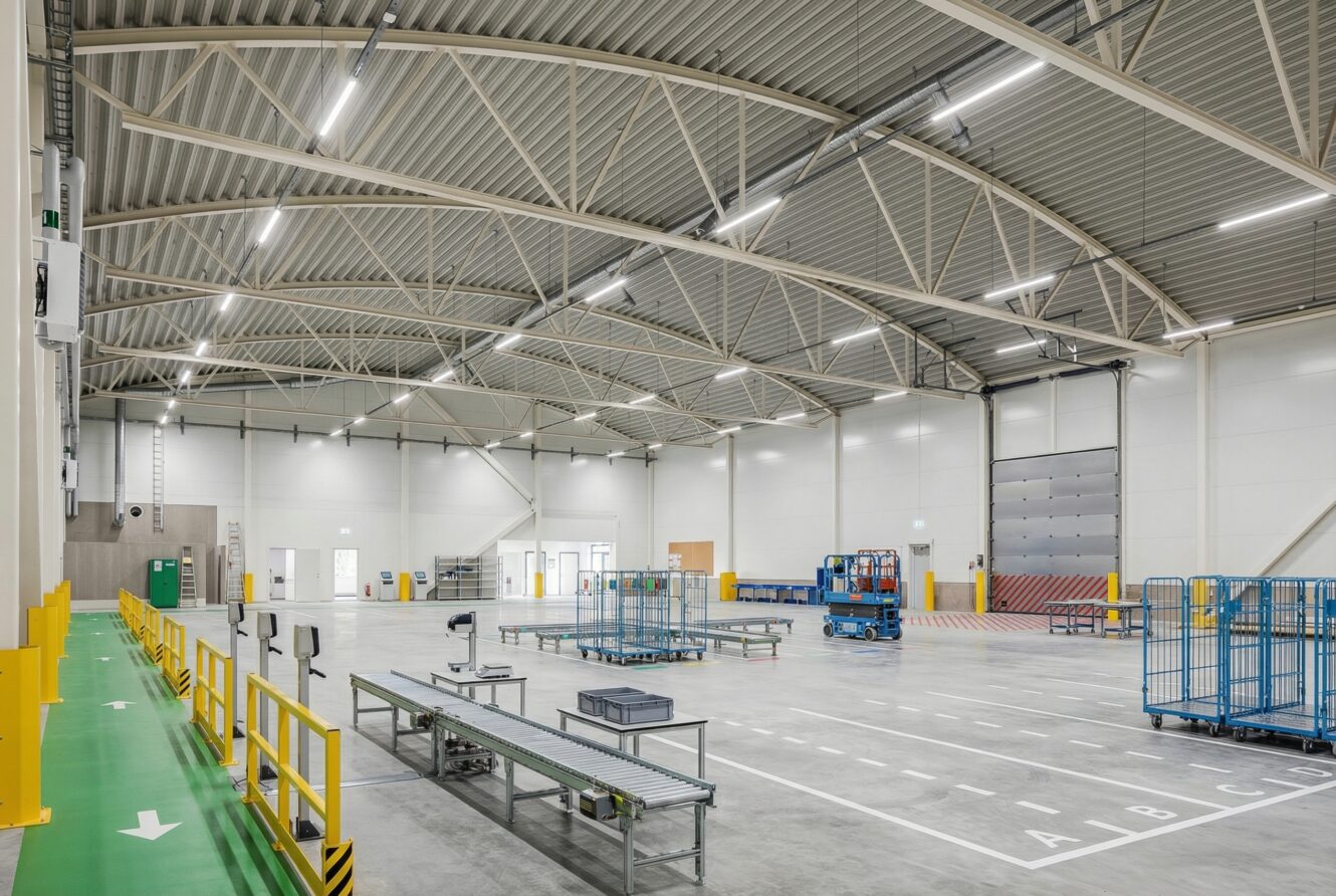 Stor tom lagerlokal med metallhyllor, transportband, lastkajer och stark takbelysning.