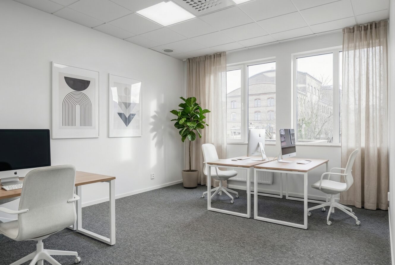 Modernt kontor med två skrivbord, datorer, vita stolar, väggkonst, en krukväxt och stora fönster med beige gardiner som släpper in naturligt ljus.