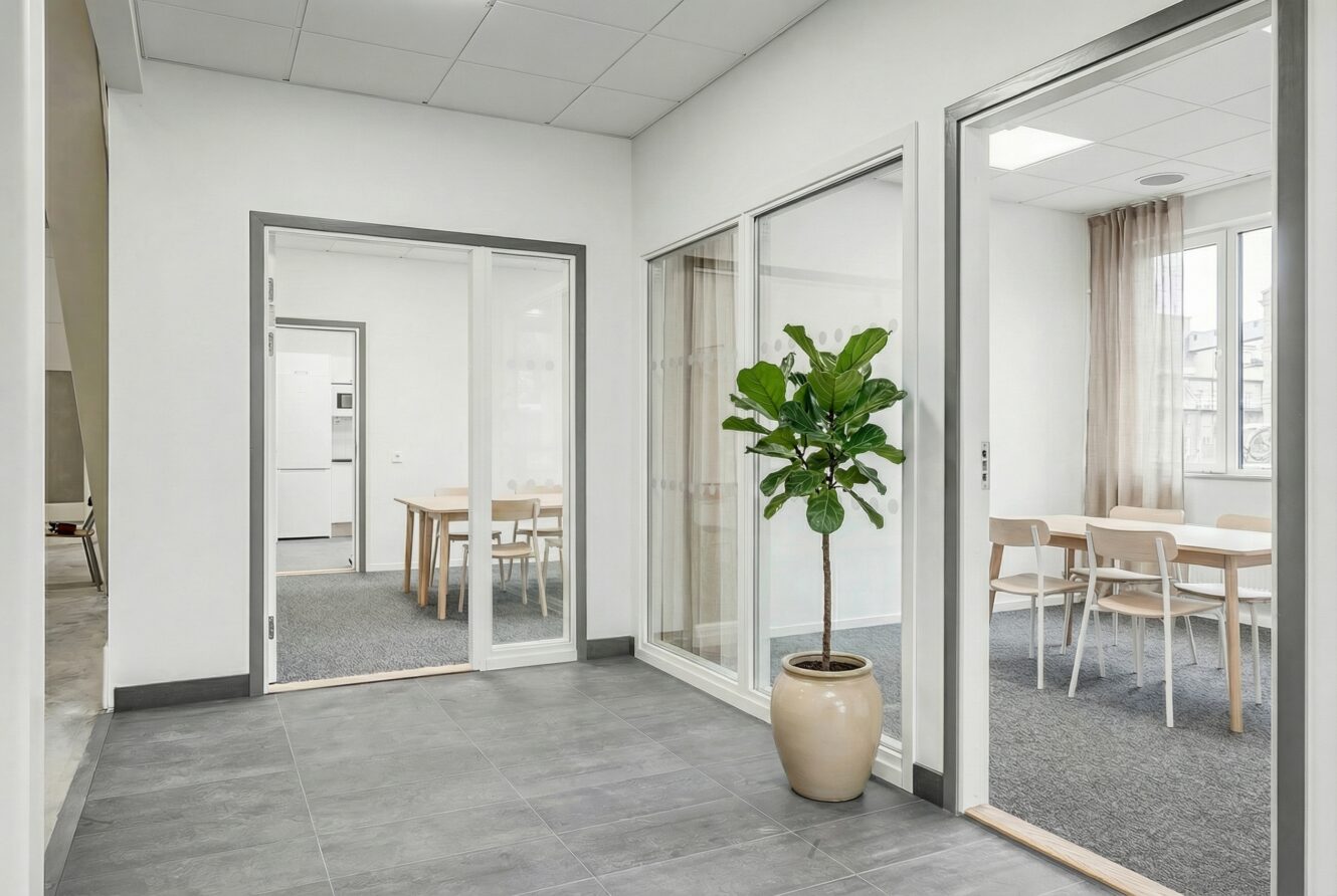Modern kontorsinredning med grått klinkergolv, en krukväxt och två mötesrum möblerade med träbord och stolar som syns genom glasdörrar.