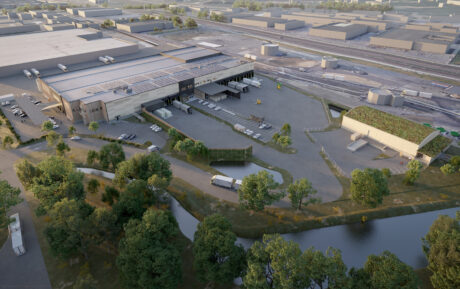 Flygfoto över en industrianläggning med lagerlokaler, lastkajer, parkerade lastbilar, grönska, en damm och omgivande vägar.