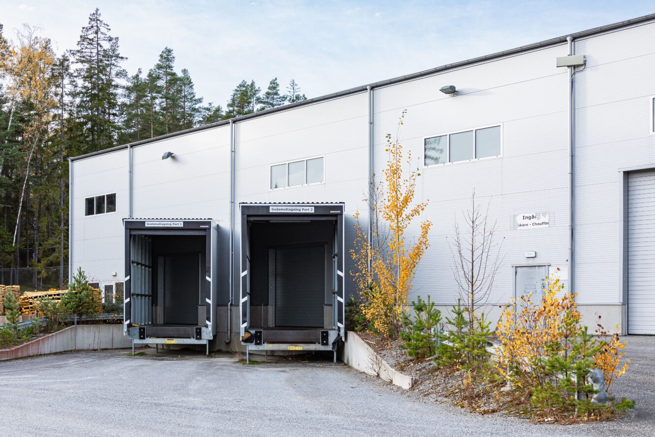 Två tomma lastkajer utanför en grå industriell lagerbyggnad, omgiven av små träd och grusmark.
