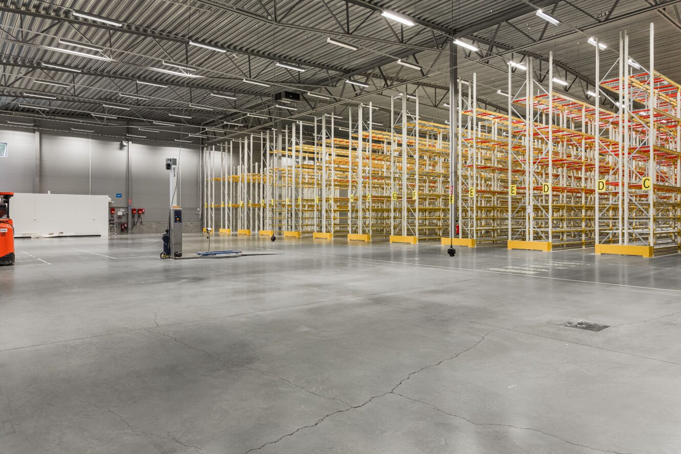 En rymlig lagerlokal med tomma industrihyllor, betonggolv, högt i tak och stark takbelysning.