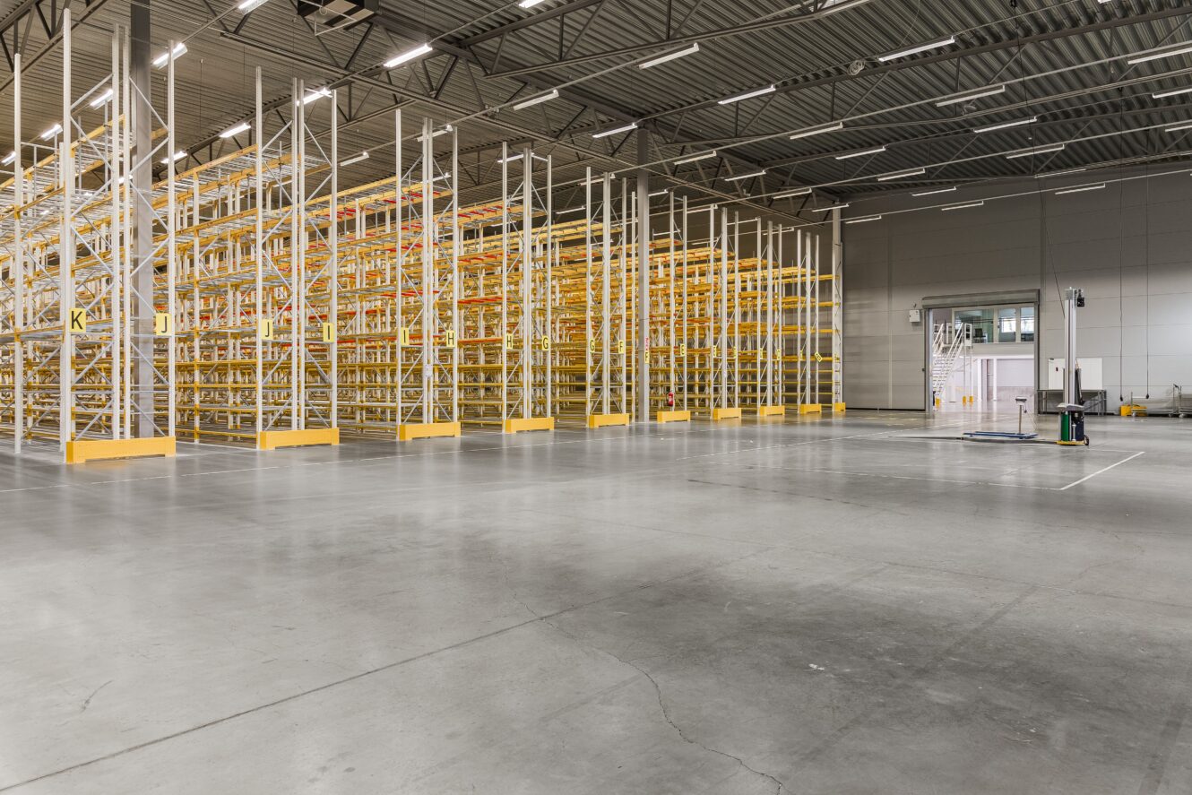 Stor tom lagerlokal med höga lagerhyllor i metall och betonggolv, upplyst av taklampor. Inga produkter eller människor är närvarande.