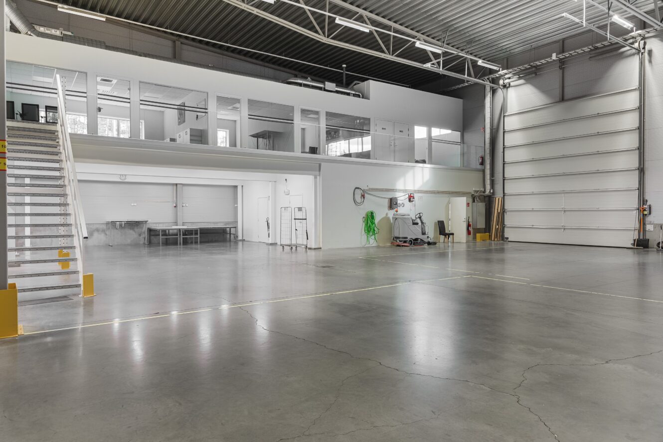 Rymlig, tom industrilokal med betonggolv, högt i tak, trappa och en övre kontorsdel med fönster. Stor garageport och viss utrustning på höger sida.