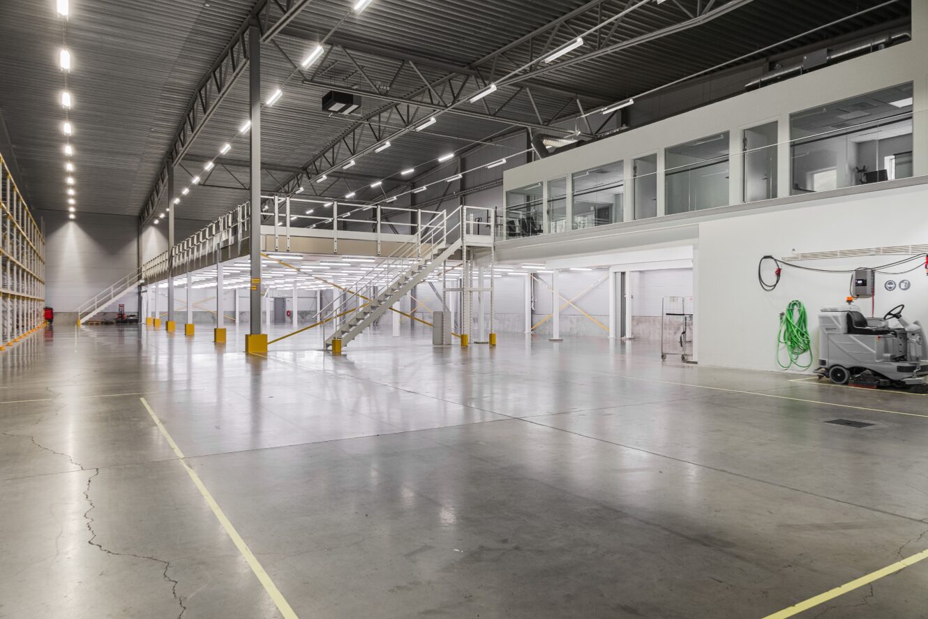 Stort, tomt industrilager med polerade betonggolv, högt i tak, takbelysning, entresol och slutna kontorsutrymmen på höger sida.