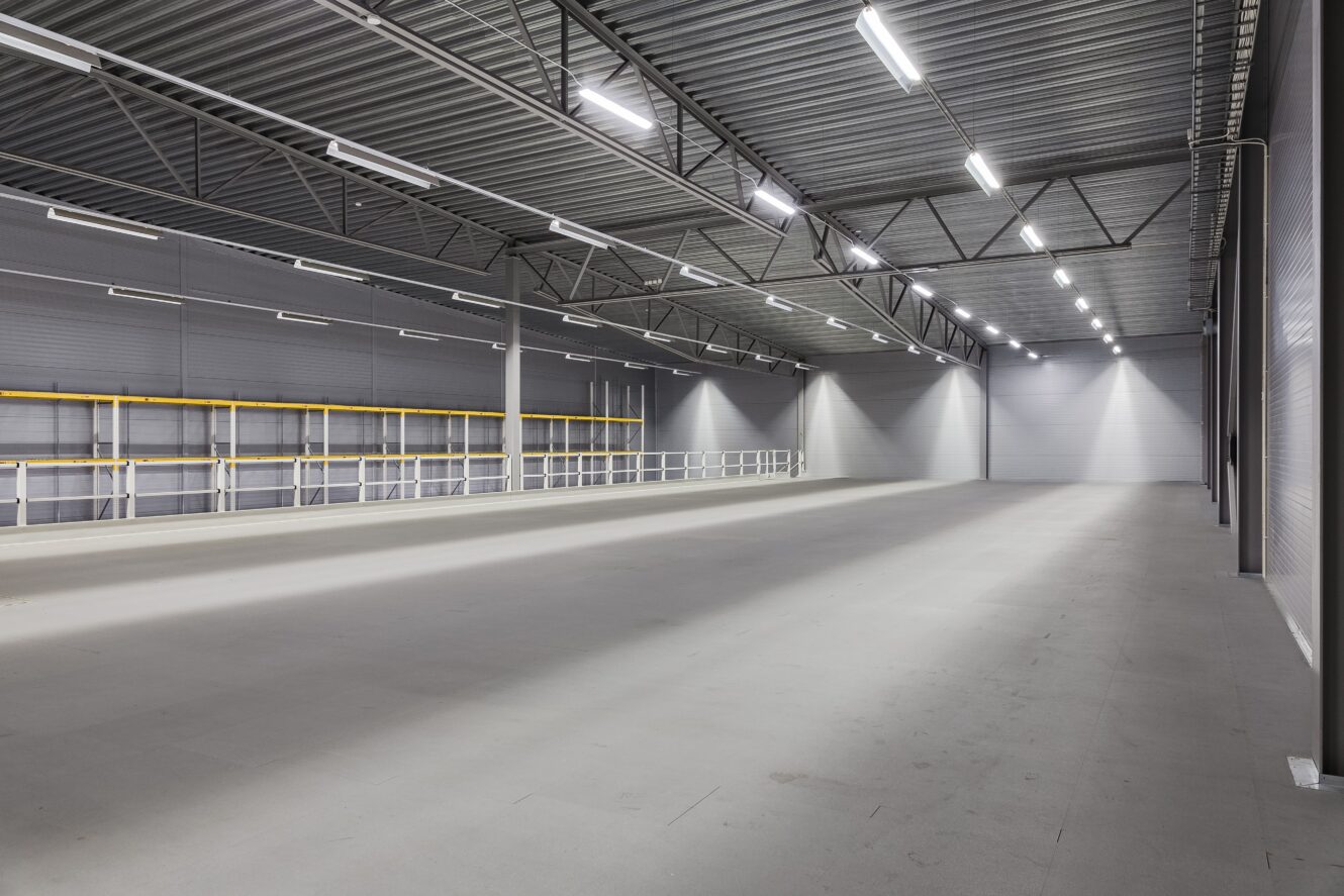 Interiör i en stor, tom lagerlokal med betonggolv, metallväggar, högt i tak och stark takbelysning.