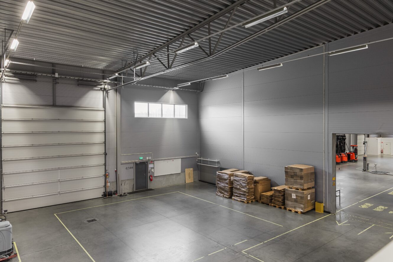 Interiör av en stor, tom lagerlokal med högt i tak, staplar av pallar och kartonger, betonggolv och industriell belysning.