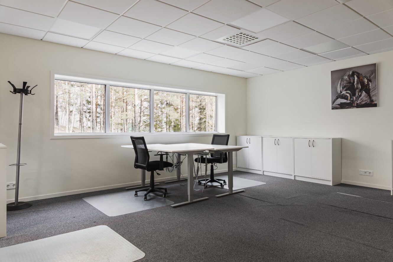 Ett modernt kontor med två stolar vid ett skrivbord, förvaringsskåp, en klädhängare och ett stort fönster med träd utanför. På väggen hänger en inramad tavla.