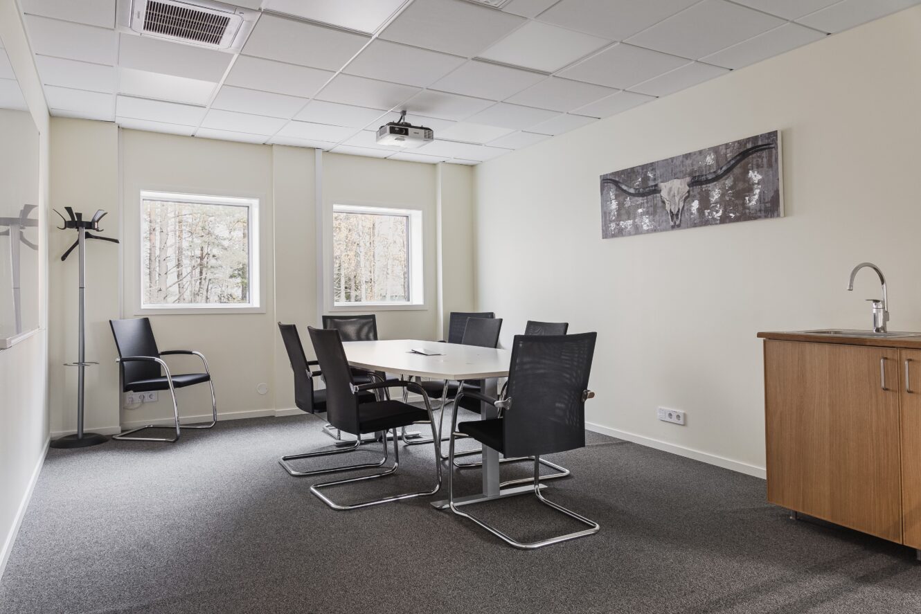 Ett litet konferensrum med ett bord, sex svarta stolar, en väggmonterad tavla, en klädhängare, två fönster och ett träskåp med handfat.