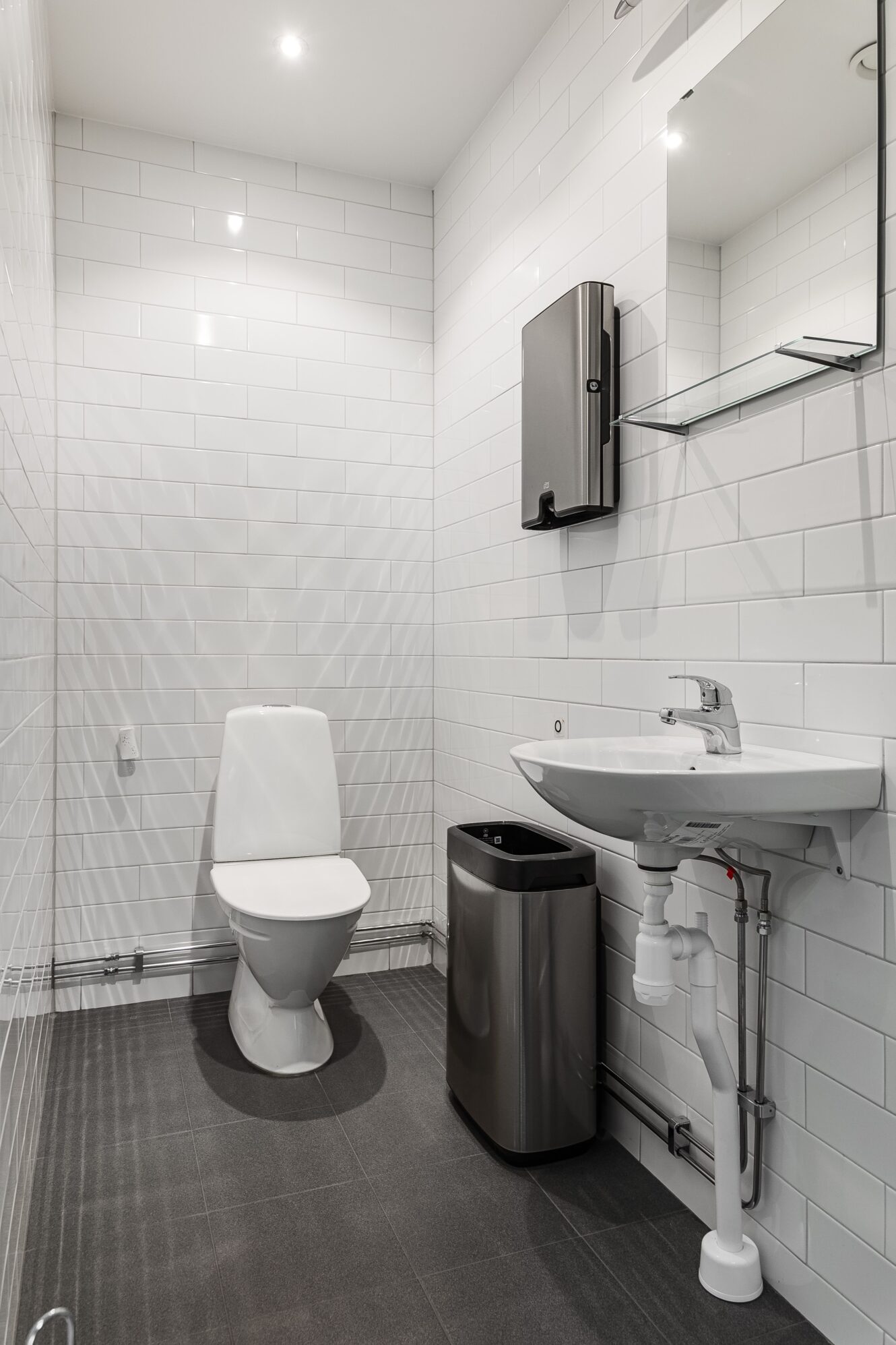 Ett modernt badrum med vitkaklade väggar, en toalett, ett vägghängt handfat, en spegel, en tvålpump och en papperskorg i rostfritt stål.
