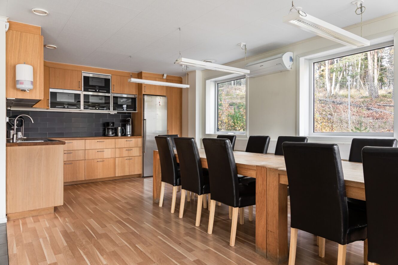 Modernt kök och matplats med ett långt träbord, svarta stolar, stora fönster, inbyggda vitvaror och skåp i ljust trä.