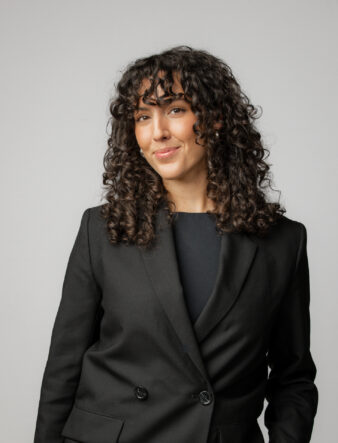 Kvinna med lockigt hår och svart kavaj står mot en ljusgrå bakgrund och tittar in i kameran med ett litet leende.