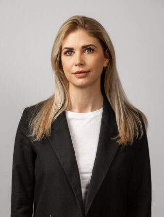 En kvinna med långt blont hår, klädd i en svart kavaj och en vit skjorta, står mot en ljusgrå bakgrund.