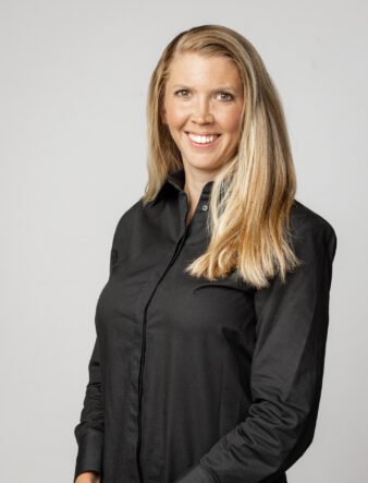 En kvinna med långt blont hår och svart skjorta med knappar står mot en ljus bakgrund och ler mot kameran.