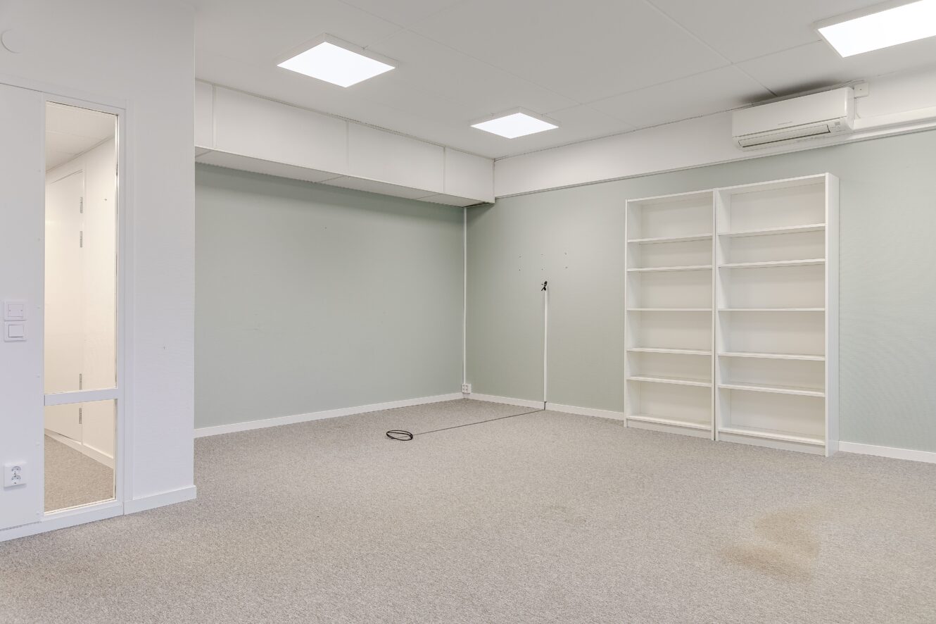 Tomt rum med ljus heltäckningsmatta, ljusgröna väggar, en vit hylla, taklampor, en luftkonditionering och ett synligt vägguttag med sladd.