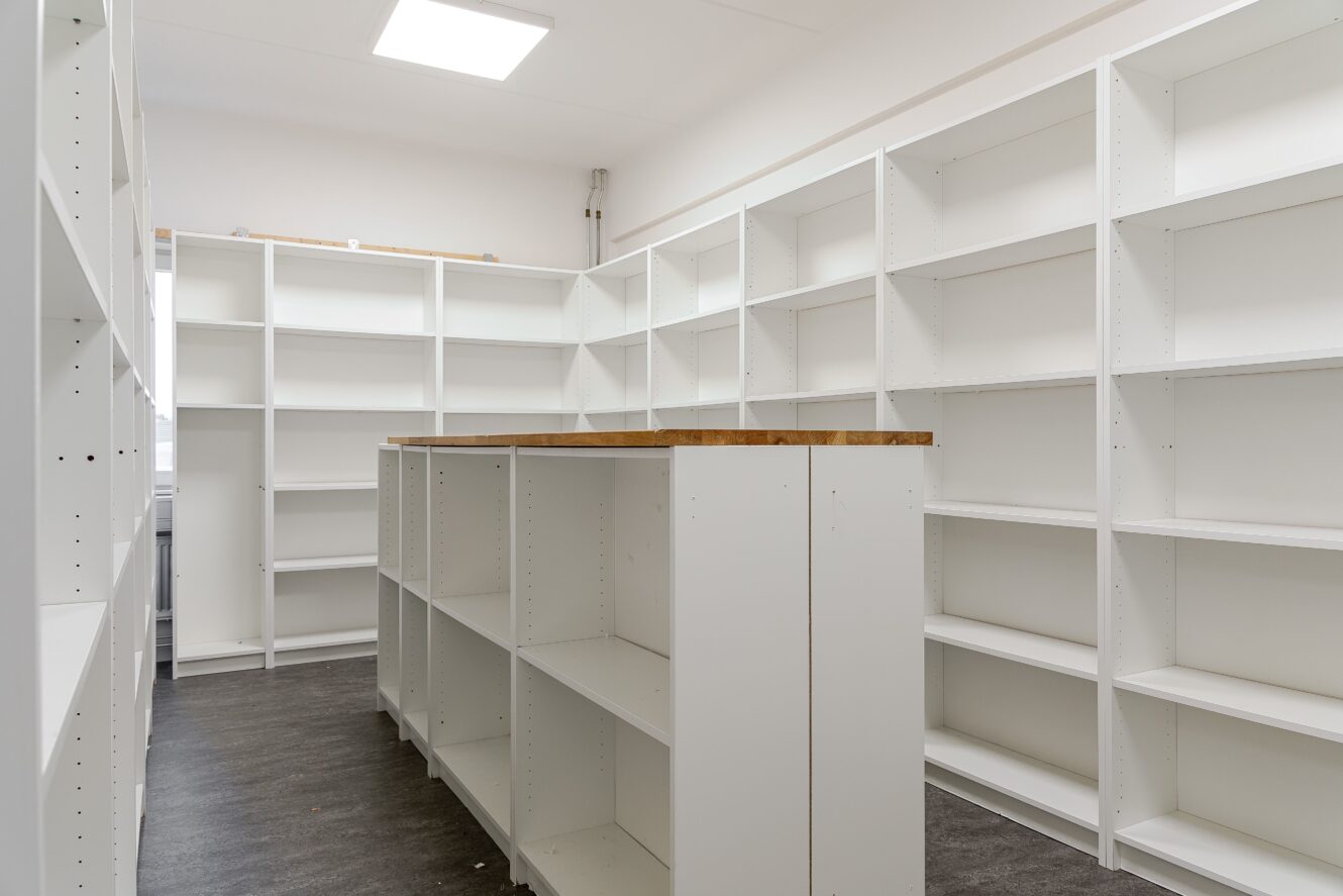 Rum med vita tomma hyllor längs väggarna och en central hylla i mitten, med ljusa taklampor och mörkt golv.