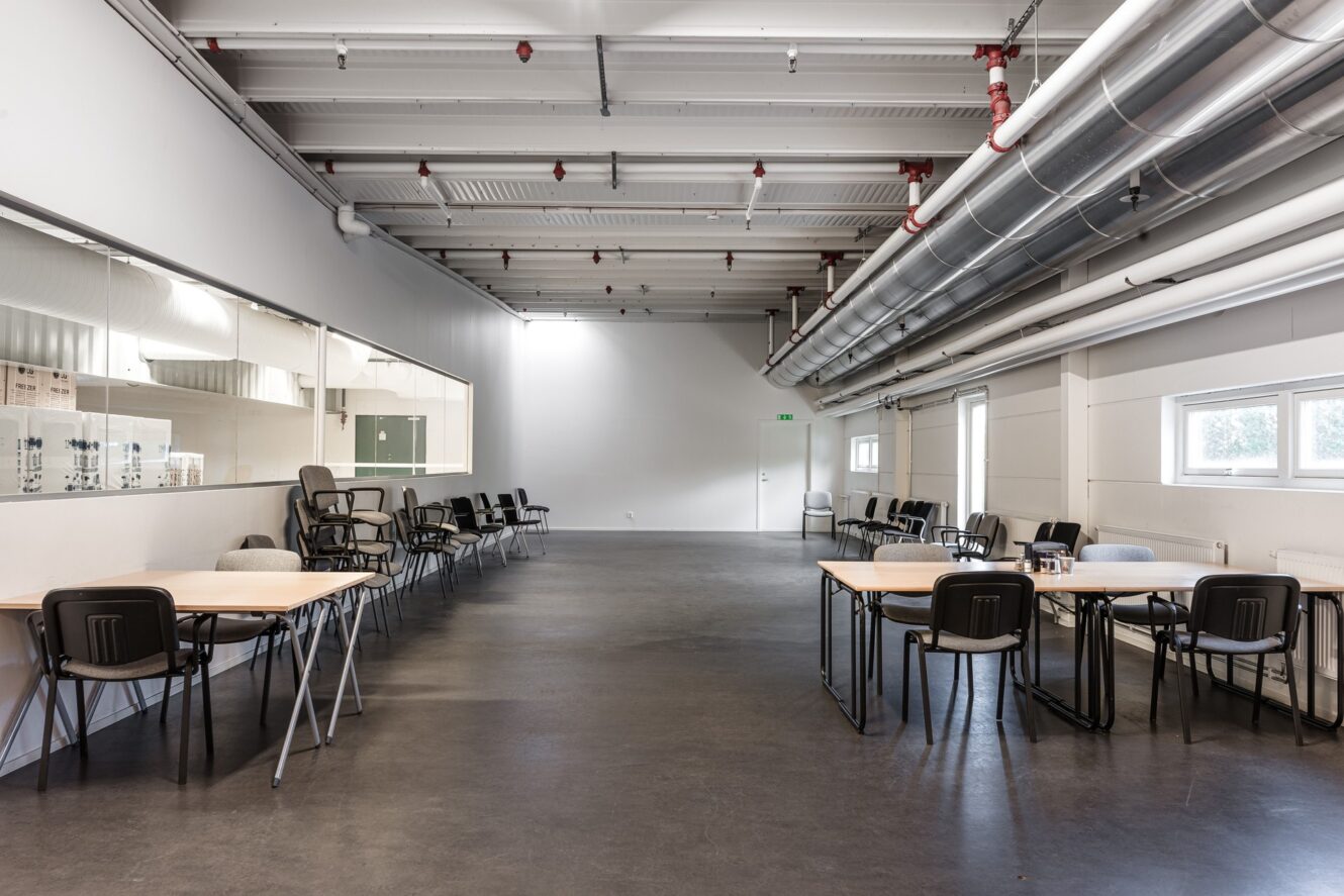 Stort, tomt rum med flera bord och stolar längs väggarna, industriellt tak och stora fönster på höger sida.
