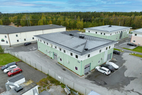 Flygfoto över en ljusgrön industribyggnad med parkerade bilar på en inhägnad tomt, omgiven av andra liknande byggnader och träd i bakgrunden.