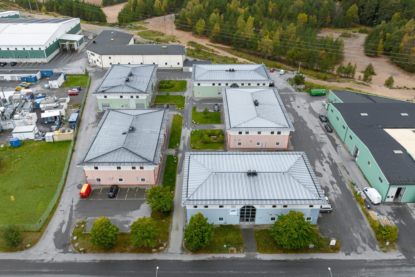 Flygfoto över flera rektangulära industribyggnader med plåttak, omgivna av parkeringsplatser, träd och grönska, belägna i närheten av ett skogsområde.