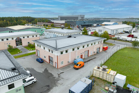 Flygfoto över ett industrikomplex med lagerbyggnader, parkerade fordon och containrar en molnig dag.