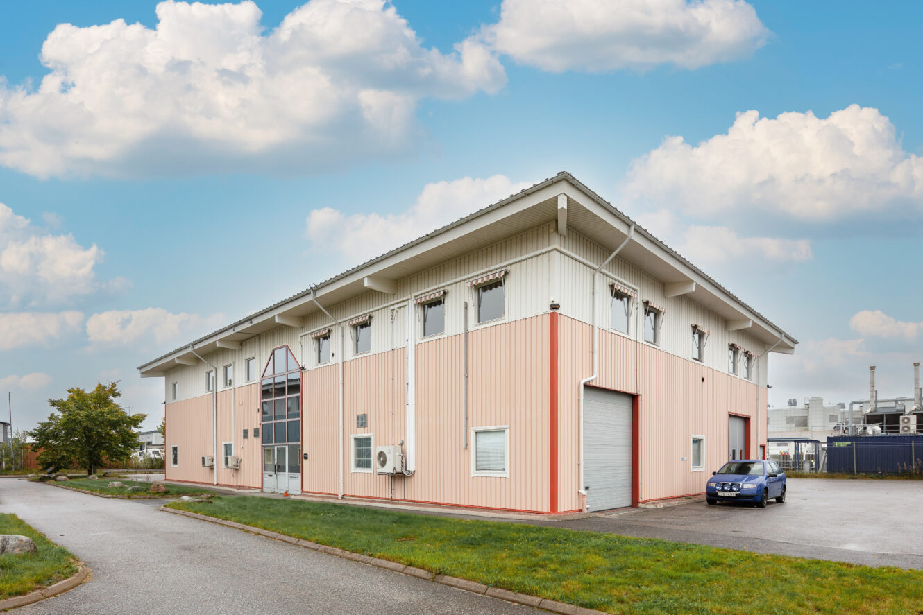 En tvåvånings industribyggnad med vita och ljusröda paneler, stora fönster och en garageport, med en blå bil parkerad i närheten under en delvis molnig himmel.