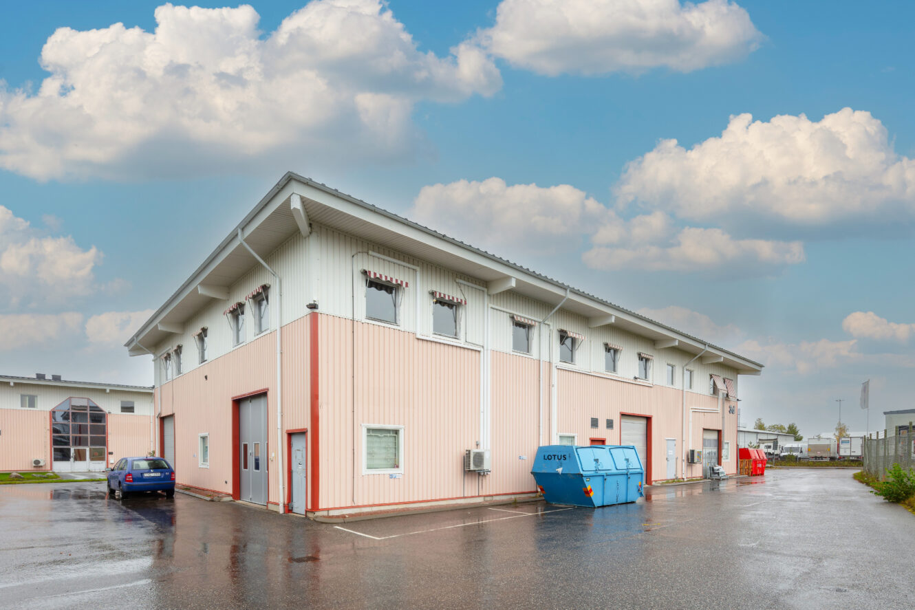 En stor, ljus industribyggnad med garageportar, en blå sopcontainer och parkerade bilar på en våt asfaltsyta under en delvis molnig himmel.