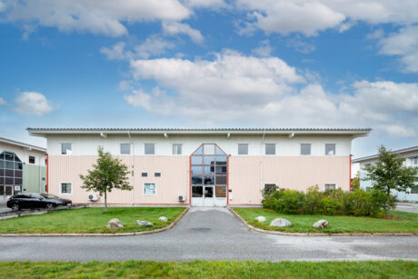 En tvåvånings ljusrosa kontorsbyggnad med en central glasentré, omgiven av gräs, buskar och en parkerad svart bil till vänster.