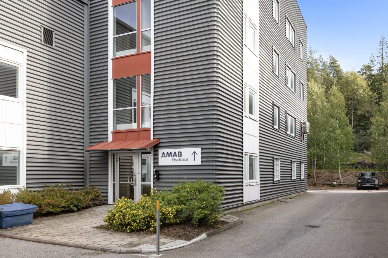 Modern grå och vit kontorsbyggnad med en skylt som visar AMAB Hydraul och en pil som pekar åt höger, omgiven av träd och en parkerad bil i bakgrunden.