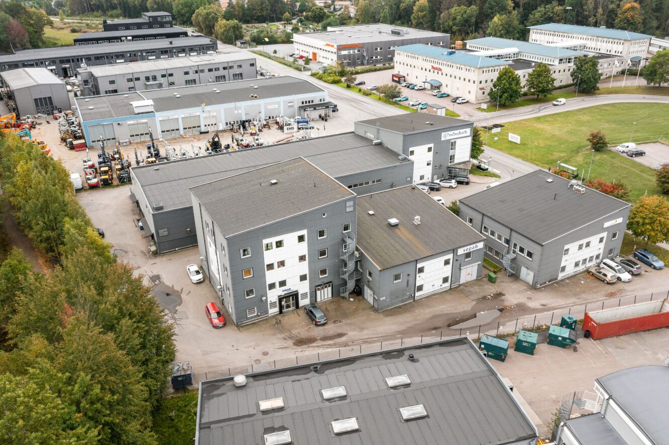 Flygfoto över ett industriområde med flera gråa kommersiella byggnader, parkerade fordon, sopcontainrar och omgivande grönska.