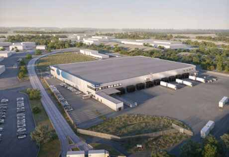 Flygfoto över en stor lagerlokal med flera lastkajer, parkerade lastbilar, intilliggande parkeringsplatser och omgivande grönska.