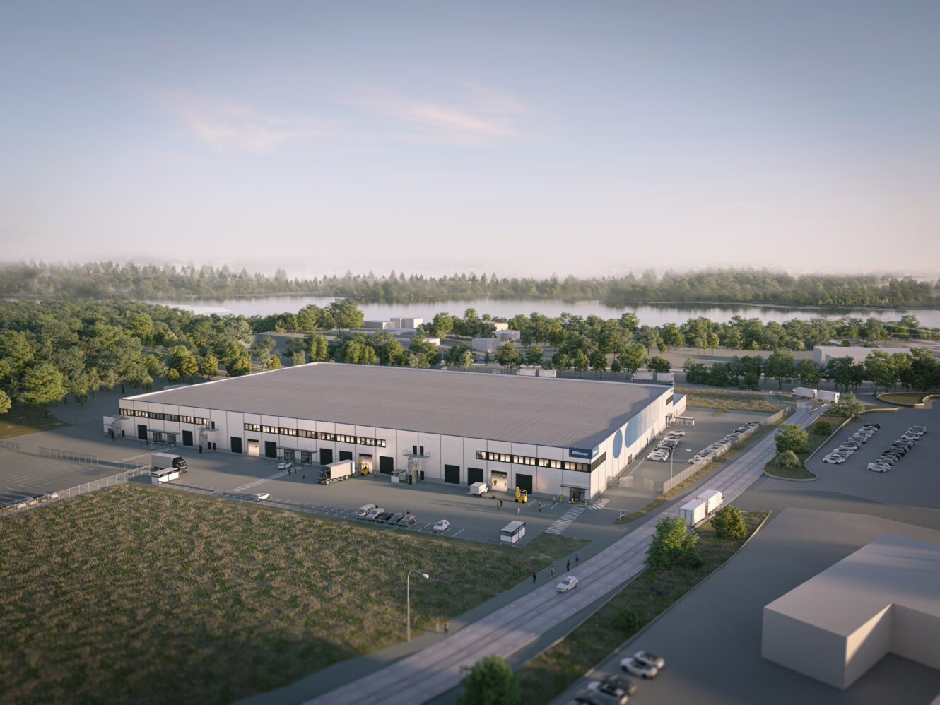 Flygfoto över ett stort, rektangulärt industrilager med lastkajer, omgivet av parkeringsplatser, vägar, träd och grönska i fjärran.