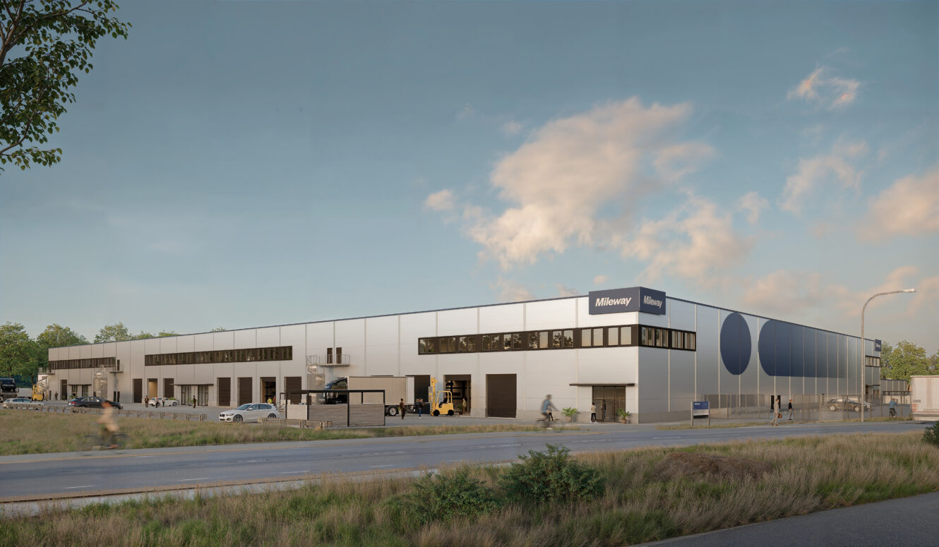 Ett stort, modernt industrilager med lastbilar, skåpbilar och cyklar framför, intill en väg med en gräsbevuxen förgrund och en delvis molnig himmel.