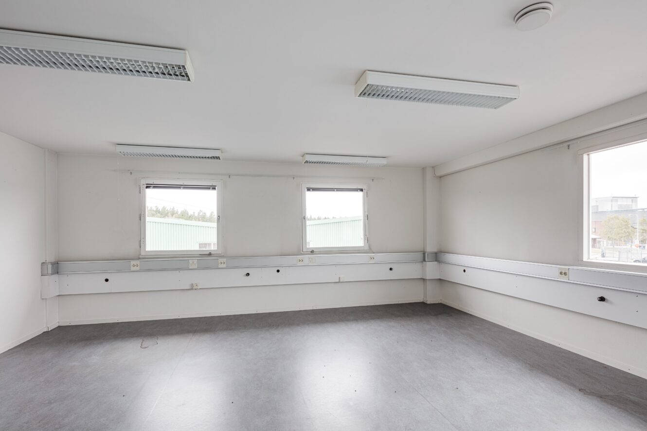 Tomt kontorsrum med tre fönster, vita väggar, taklampor, grått golv och eluttag längs väggarna.