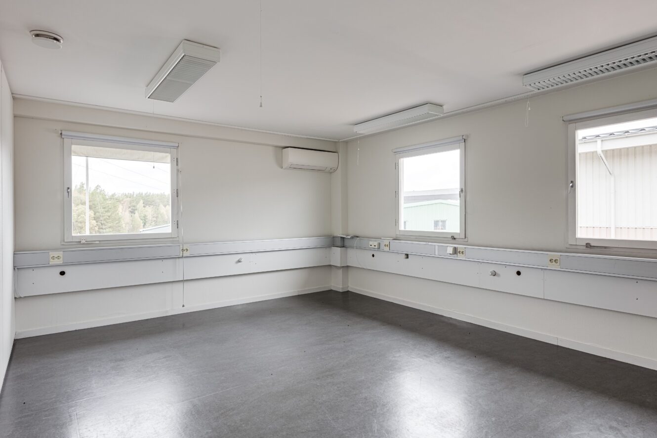 Tomt, omöblerat rum med tre fönster, vita väggar, grått golv, väggmonterade eluttag och taklampor. En luftkonditioneringsenhet finns ovanför ett fönster.