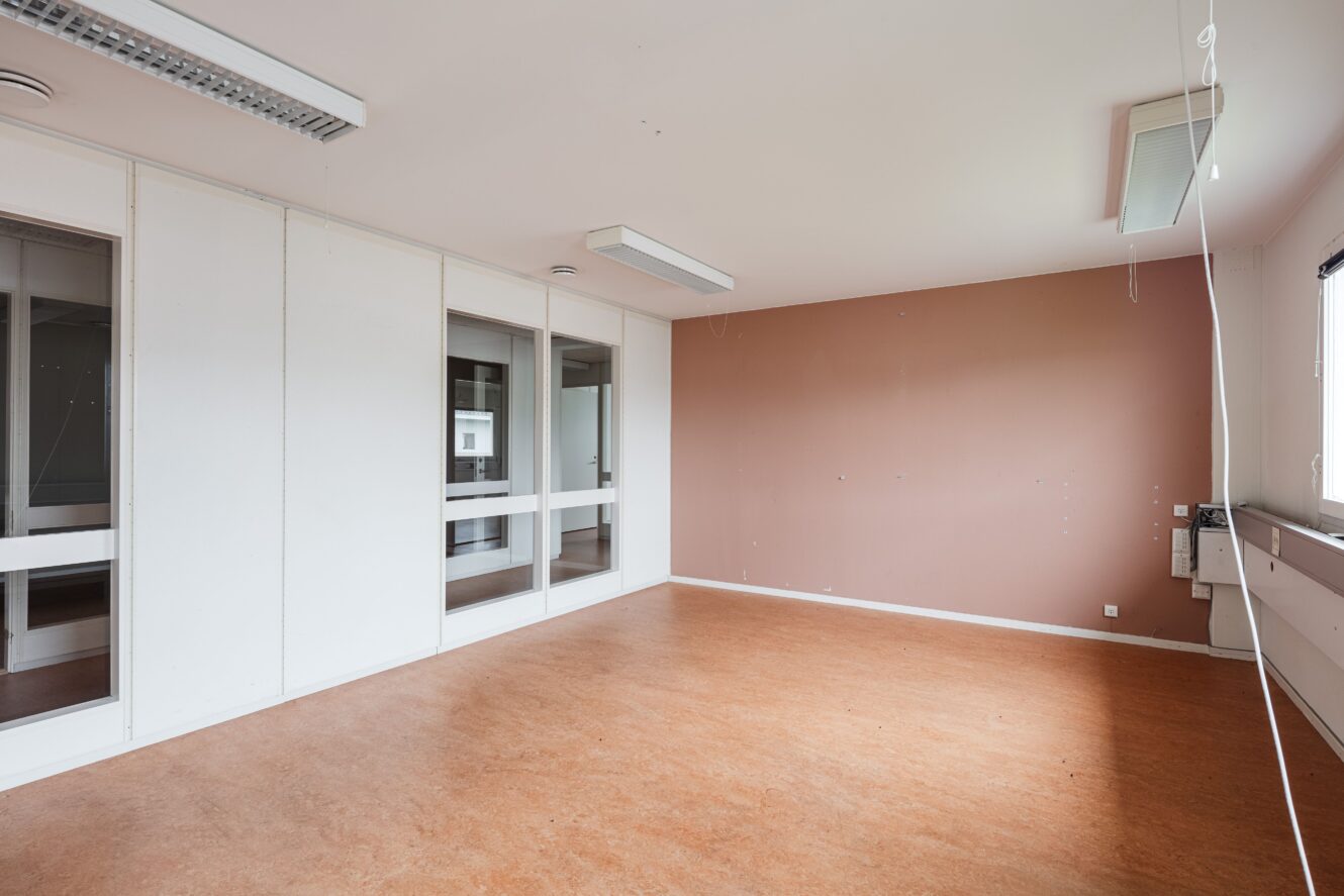 Ett tomt kontorsrum med brunt golv, en brun accentvägg, vita väggar med glaspaneler, takbelysning och ett fönster på höger sida.