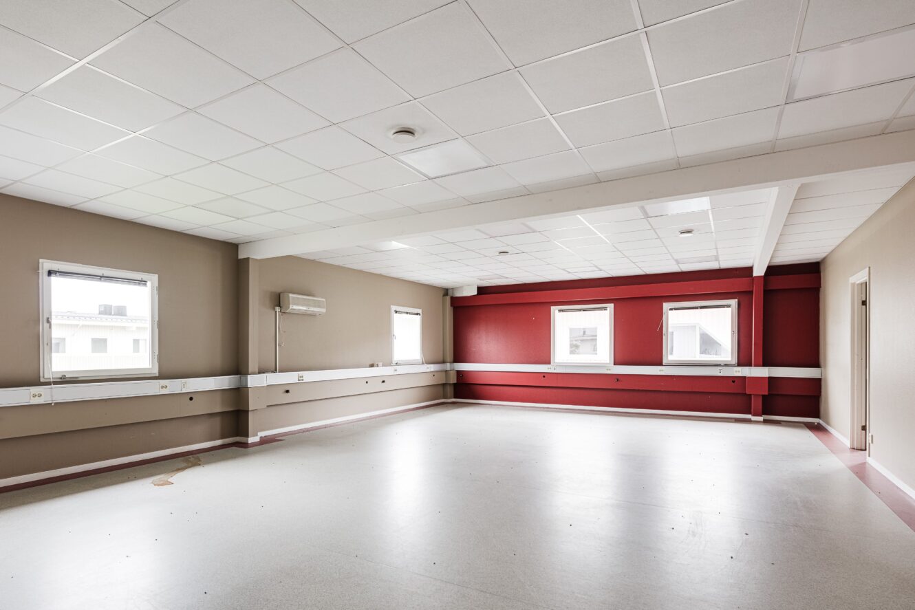 Tomt, omöblerat rum med vitt tegeltak, beige och röda väggar, tre fönster och väggmonterade eluttag längs med väggarna.