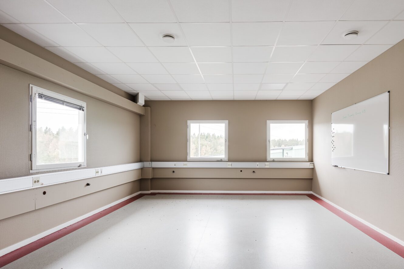 Ett tomt rum med beige väggar, tre fönster, en whiteboard på den högra väggen och takplattor. Golvet är ljust med en röd bård längs kanterna.